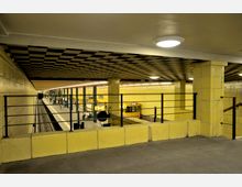 Untergrundbahnhof mit gelb gefliesten Wänden und schwarzem Schachbrettmuster an der Decke. Ein Bahnsteig mit wartenden Personen, Schienen, sowie ein gelber Zug sind sichtbar, umgeben von metallenen Geländern und Stützpfeilern.