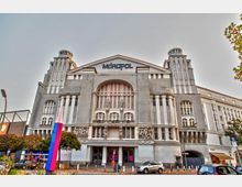 Fassade eines großen historischen Gebäudes mit der Aufschrift „Metropol“ am Giebel. Das graue Gebäude hat architektonische Verzierungen, hohe Fenster, zwei Türme und skulpturale Elemente an der Front.