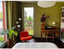 Gemütliches Schlafzimmer mit olivgrünen Wänden, großem Papier-Deckenlampenschirm und dunklem Holzboden; links ein roter Sessel neben einer hohen Stehlampe und einer großen Topfpflanze. Rechts steht ein Holzschreibtisch mit Stuhl, Tischlampe und Blumenvase, daneben ein Bücherregal; durch bodentiefe Fenster und eine Glastür mit Vorhängen blickt man auf einen Balkon und viel Grün im Außenbereich.