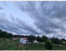 Ein ländliches Grundstück mit einer weißen Einfamilienhaus mit rotem Dach inmitten einer grünen Wiese und jungen Bäumen. Im Hintergrund ist ein bewölkter Himmel zu sehen, und am Rand sind weitere kleine Gebäude sowie eine Antenne erkennbar.