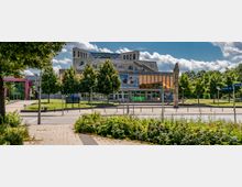 Außenansicht der Uckermärkischen Bühnen Schwedt, einem modernen Theatergebäude mit großer Glasfassade und dahinterliegendem Terrassenaufbau. Im Vordergrund befinden sich eine Straßenkreuzung, grüne Bepflanzung sowie Bäume entlang der Fassade.