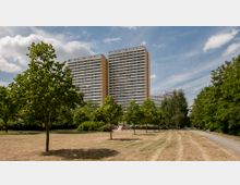 Hochhäuser mit hellen Fassaden und orangefarbenen Elementen ragen hinter einem Parkbereich mit Wiese, vereinzelten Bäumen und einem kleinen Spielplatz hervor. Der Himmel ist leicht bewölkt, und ein gepflasterter Weg verläuft am rechten Bildrand.