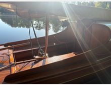 Blick ins Innere eines hölzernen Segelboots mit Mast, Tauwerk und aufgerolltem Segel, das auf einem ruhigen See liegt. Im Hintergrund spiegeln sich Bäume am Ufer im Wasser, während Sonnenstrahlen über das Boot fallen.