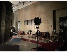 Ein modern eingerichteter Raum mit industriellem Stil, gekennzeichnet durch Betonwände, hängende Deckenlampen und ein Filmbeleuchtungsgerät im Vordergrund. Im Hintergrund befinden sich Ledersessel, ein Couchtisch mit einer Vintage-Tischlampe und mehrere Schwarz-Weiß-Bilder an der Wand.