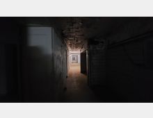 Langer, dunkler Flur mit abblätternder Farbe an den Wänden und einer freiliegenden Decke mit Kabeln. Auf beiden Seiten befinden sich Türen und der Boden ist mit Fliesen bedeckt.