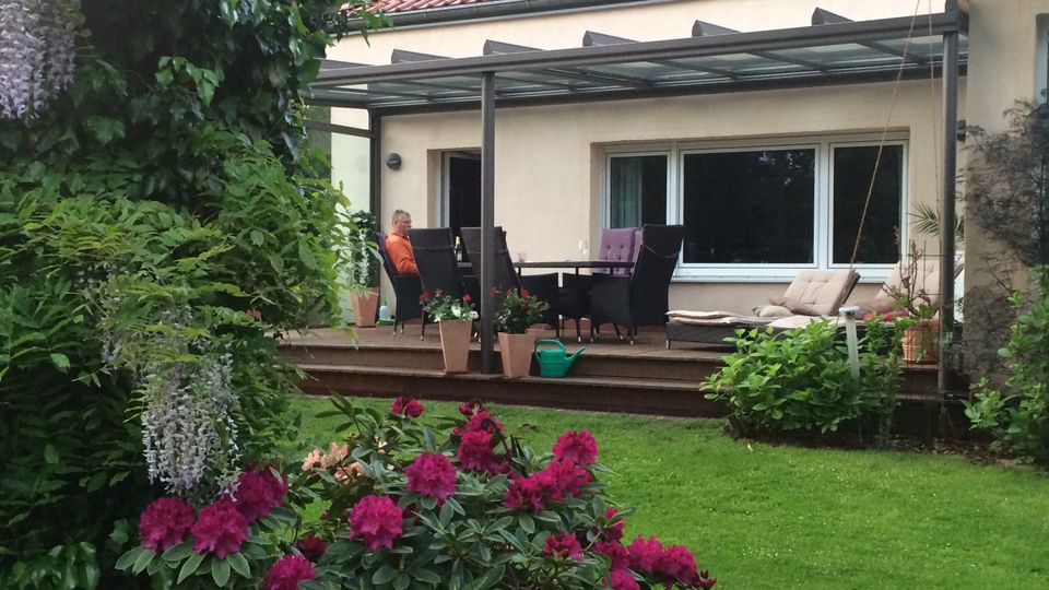 Terrasse an einem Wohnhaus mit rotem Ziegeldach, überdacht von einer Glas- und Metallkonstruktion, davor ein Esstisch mit Stühlen und Pflanzkübeln. Im Vordergrund liegt ein grüner Garten mit Rasen und blühenden Sträuchern sowie dichtem Baumbestand am Rand.