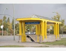 Gelber, pavillonartiger Eingang zum U-Bahnhof Haselhorst mit blauem Schild; Zugang zur Treppe ist sichtbar. Im Hintergrund eine Straße, Verkehrsschilder sowie Bäume in einer herbstlichen Umgebung.