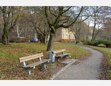 Ein kleiner Park mit Laubbäumen im Herbst, zwei Holzbänken neben einem befestigten Gehweg und einer Wiese mit fallendem Laub. Im Hintergrund sind Wohngebäude mit braunen Fassaden und weißen Fensterrahmen zu sehen.