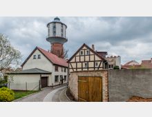 Straßenszene in einem Dorf mit einem historischen Wasserturm im Hintergrund, Fachwerkhäusern und Wohngebäuden mit roten Ziegeldächern. Im Vordergrund ist eine gepflasterte Straße sowie eine Mauer mit einem Holztor zu sehen.