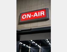 Leuchtendes rotes Schild mit der Aufschrift „ON-AIR“ in weißen Buchstaben, montiert an einer Betondecke über einer Tür. Im Hintergrund sind Teile eines dunklen Raums mit modernen Deckenleuchten sichtbar.