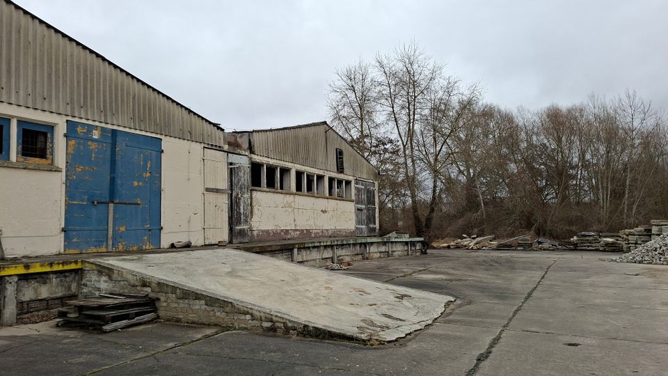 Verlassene Lager- oder Industriehalle mit Wellblechdach, abblätternden blauen Toren und einer Betonrampe zu einer Laderampe. Davor liegt ein großer, rissiger Betonhof mit Materialstapeln am Rand und kahlen Bäumen im Hintergrund.