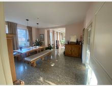 Esszimmer mit Holzmöbeln, darunter ein großer Tisch und Bänke, auf einem glänzenden Steinboden. Der Raum hat große Fenster mit beigen Vorhängen, eine Kommode an der Wand und zahlreiche Pflanzen sowie Dekorationen.