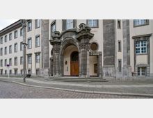 Historisches Gebäude mit einer verzierten Eingangsfassade, bestehend aus massiven Steinsäulen, Rundbogenportal und detailliertem Dekor. Der Vorplatz ist mit Kopfsteinpflaster bedeckt, flankiert von einem Gehweg und einer angrenzenden Straße.