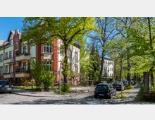 Straßenszene mit Altbauten aus rotem Backstein und hellen Fassaden, umgeben von Bäumen mit frischem, grünem Laub. Der gepflasterte Gehweg und parkende Autos fügen sich in eine ruhige Wohngegend ein.