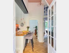 Helle, moderne Wohnung mit Blick auf eine offene Küche mit Holztheke und drei gepolsterten Barhockern, hölzernem Fischgrätboden, weißen Wänden und einer rosa Decke. Im Hintergrund befinden sich Zimmerpflanzen, ein Essbereich mit Glastüren und Grünelemente draußen.