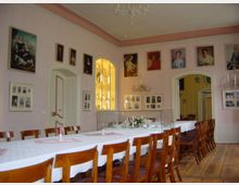 Ein historischer Speiseraum mit einer langen, gedeckten Tafel und Holzstühlen. Die rosa Wände sind mit Gemälden und Fotografien dekoriert, und in einer beleuchteten Vitrine werden Porzellanstücke präsentiert.