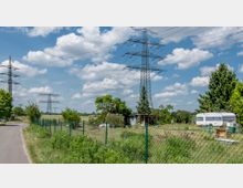 Ländliche Szene mit Strommasten und einer kleinen Gartenparzelle, die durch einen Maschendrahtzaun abgegrenzt ist. Im Garten befinden sich ein Wohnwagen, ein kleines Holzhaus und verschiedene Bäume, umgeben von einer offenen Feldlandschaft unter einem teilweise bewölkten Himmel.