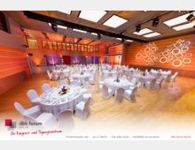 Ein großer, modern gestalteter Veranstaltungssaal mit Holzfußboden, rötlich beleuchteten Wänden und einer dekorativen Lichtprojektion in Form von Kreisen und Quadraten. Der Raum ist mit mehreren runden Tischen gedeckt, die weiße Tischdecken, Stuhlhussen und Blumenarrangements als Mittelstücke haben.