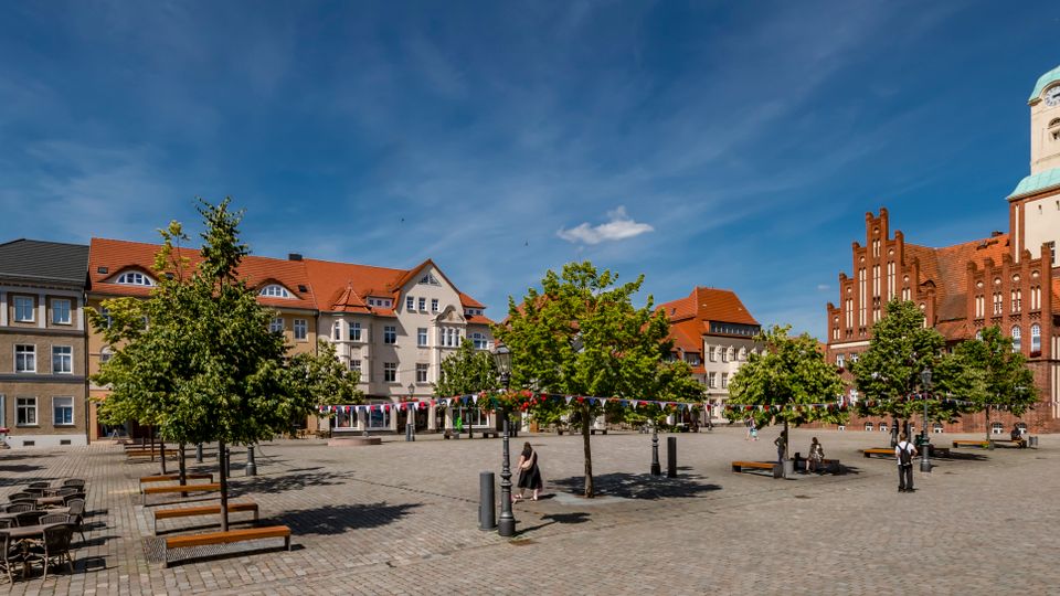 Großer gepflasterter Marktplatz mit Bänken, kleinen Bäumen, Laternen und einer Reihe von Wimpelgirlanden über dem Platz. Am Rand stehen mehrstöckige Wohn- und Geschäftshäuser sowie eine große Backsteinkirche mit hohem Turm und kupfergrünem Dach.