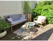 Kleine Terrasse mit Steinboden, möbliert mit einer Holzbank, einem Holzsessel und zwei runden Beistelltischen. Umgeben von grünen Pflanzen und Sträuchern, mit einem Bambuskübel als Dekoration.