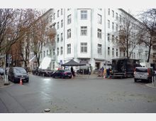 Straßenszene an einer Ecke mit einem weißen mehrstöckigen Wohngebäude, vor dem ein Film- oder Fotoshooting stattfindet. Fahrzeuge, Ausrüstungswagen und Pavillons mit technischem Equipment stehen entlang der Straße; kahle Bäume und geparkte Autos säumen den Straßenrand.