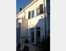 Zweistöckiges Wohnhaus mit heller Fassade und schwarzen Fensterläden, gezeigt von einer Terrasse aus. Vor der Fassade befinden sich Pflanzenkübel, Outdoor-Möbel und ein zusammengerollter Gartenschlauch.