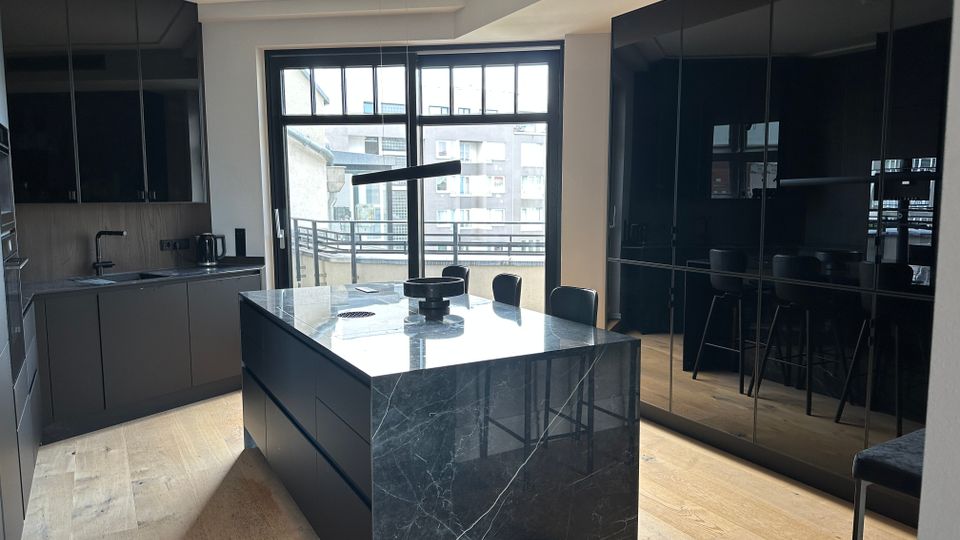Moderne Wohnküche mit großer Kochinsel aus dunkel marmorierter Steinplatte und umlaufenden mattschwarzen Schrankfronten. Eine breite Fensterfront mit Balkontür bringt Tageslicht hinein; rechts sind hochglänzende, spiegelnde Schranktüren und Holzfußboden zu sehen.