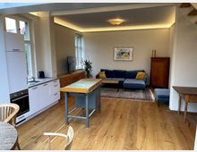 Wohnbereich mit offener Küche, Holzboden und gemütlicher Einrichtung. Links befindet sich eine weiße Küchenzeile, während rechts eine blaue Sofa-Lounge mit Teppich und Couchtisch zu sehen ist; im Hintergrund steht ein Holzschrank und ein Fernseher.