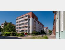 Mehrstöckiges Wohngebäude mit Balkonen und rot-weißer Fassade an einer ruhigen Straße mit Gehwegen, Bäumen und parkenden Fahrrädern. Im Vordergrund sind Grünflächen und Büsche sichtbar, im Hintergrund weitere Wohnhäuser.