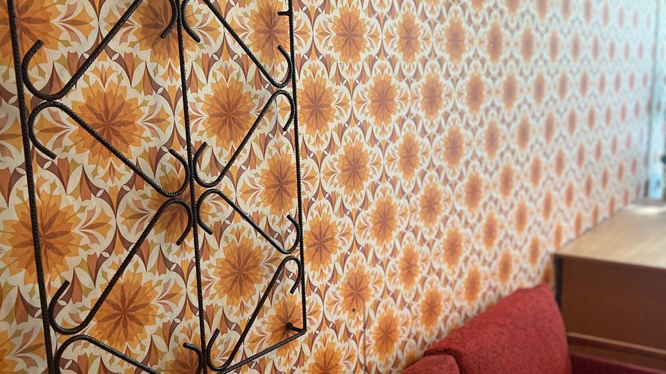 Innenraum mit einer langen Wand in orange-brauner, geometrisch-floraler Retro-Tapete und einem schwarzen Metall-Garderobenständer mit Haken. Im Vordergrund steht ein rotes Polstersofa, am rechten Rand ist ein helles Holzregal zu sehen.