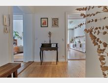 Flur in einer Wohnung mit Holzboden, einem schmalen dunklen Tisch mit dekorativer Vase und Bildern an der Wand, verbunden mit Ausblicken in ein Wohnzimmer links und eine Küche rechts mit weißen Einbauschränken und hoher Decke.