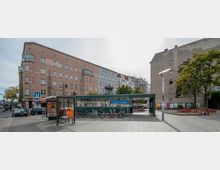 Eingangsbereich des U-Bahnhofs Yorckstraße in Berlin, umgeben von mehrstöckigen Wohngebäuden. Im Vordergrund stehen Fahrräder vor einem Wartehäuschen und einer Bushaltestelle; daneben Bäume und ein kleiner Platz mit Sitzmöglichkeiten.