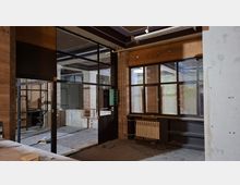 Verlassener Innenraum eines ehemaligen Büro- oder Werkstattbereichs mit Glas- und Metalltrennwänden, hohen Fenstern und sichtbaren Deckenbalken. Abgeblätterte Wände, freiliegende Leitungen, ein Heizkörper unter dem Fenster und Bauschutt am Boden prägen die Umgebung.