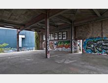Verlassene Industriehalle mit offenen Stahlträgern und Betonboden, deren Wände mit Graffiti bedeckt sind. Im Hintergrund sind eine blau gestrichene Außenwand und überwuchertes Grün sichtbar.