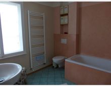 Badezimmer mit hellen Wänden in Beige und Rosa, einer Badewanne, einem Wand-WC und einem weißen Waschbecken. Neben einem Fenster befindet sich ein weißer Handtuchheizkörper, und der Boden ist mit blauen Fliesen ausgelegt.