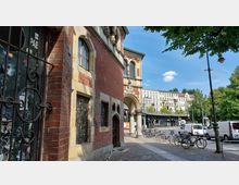 Straßenszene mit einem historischen Backsteingebäude im Vordergrund, das mit vergitterten Fenstern und Sandsteindetails gestaltet ist. Im Hintergrund sind ein erhöhtes Bahnviadukt, ein mehrstöckiges Wohngebäude und parkende Fahrräder sowie Autos auf einer breiten Straße zu sehen, umgeben von Bäumen und Straßenlaternen.