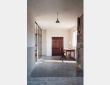 Minimalistisch eingerichteter Raum mit Betonboden, weißen Wänden und einer Holztür in der Mitte. Links ist eine Wand mit großen Glas- und Metallrahmen, rechts ein freiliegender Ziegelabschnitt, sowie ein Holztisch und ein Drehstuhl vor einem Fenster.