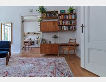 Wohnzimmer mit einer hellen Einrichtung und einem Holzfußboden, einem gemusterten Teppich und einer Bücherwand, die einen Schreibtisch integriert. Im Hintergrund ist durch eine Tür eine Küche mit Esstisch und Regalen sichtbar, die von Pflanzen und Haushaltsgegenständen dekoriert ist.