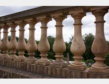 Steinbalustrade mit massiv geformten Balustern, die auf einem Ziegelsteinfundament stehen; im Hintergrund ist eine bewaldete Landschaft unter bewölktem Himmel sichtbar.