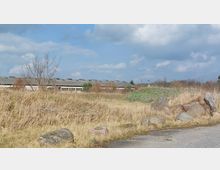 Eine flache, graue Industriehalle inmitten einer überwucherten Wiesenlandschaft mit dichtem, trockenem Gras, einigen Büschen und kleinen Bäumen. Im Vordergrund befinden sich große Steine und eine asphaltierte Fläche, während am Himmel vereinzelte Wolken zu sehen sind.