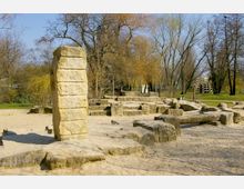 Spielplatz mit sandigem Boden und großen, unregelmäßig geformten Steinstrukturen, darunter ein hoher rechteckiger Steinblock. Im Hintergrund sind Bäume ohne Laub sowie eine Wiese zu sehen.
