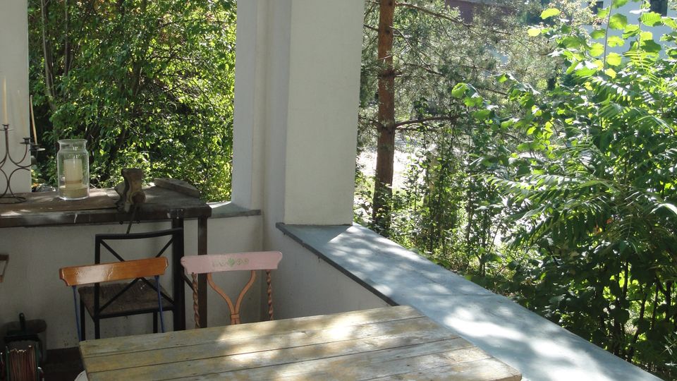 Überdachte Veranda mit weißen Rundbögen und hängender Laterne, ausgestattet mit einem kleinen Holztisch und Stühlen auf Fliesenboden. Seitlich öffnet sich der Blick ins dichte Grün von Bäumen und Sträuchern, mit einem niedrigen Geländer entlang der Kante.