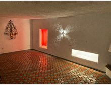 Innenraum mit weiß verputzten Wänden, buntem Mosaikboden und zwei Wandnischen, eine rot beleuchtet und die andere weiß. An der Wand hängt ein dekoratives Metallornament mit Kerzenhalter.