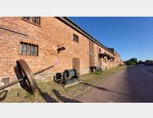 Das Bild zeigt ein langes Backsteingebäude, vermutlich ein historisches Lagerhaus, mit kleinen Fenstern, Holztüren und einem Schild mit der Aufschrift „Lokomuseum“. Im Vordergrund befinden sich eine alte Maschinenteile und gepflasterter Boden, während rechts eine breite gepflasterte Straße und im Hintergrund Bäume und parkende Autos sichtbar sind.