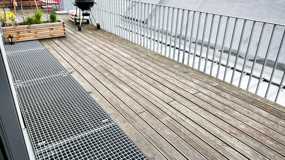 Holzterrasse mit Geländer, ausgestattet mit einem Holztisch, Stühlen, einem Grill und einem bepflanzten Blumenkasten; im Hintergrund sind umliegende Gebäude und Bäume zu sehen.