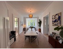 Helles Esszimmer in einem Altbau mit weiß gestrichenen Wänden, hohem Stuck an der Decke und einem großen, rechteckigen Esstisch in der Raummitte. Im Hintergrund sind ein großes Fenster mit Blick ins Grüne sowie weitere Türen und Dekorationselemente wie Pflanzen, Bilder, eine Kommode und ein Kronleuchter zu sehen.