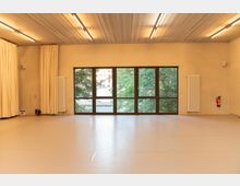 Ein heller Raum mit großen Fenstern, die nach draußen auf Bäume und ein Gebäude blicken. Der Raum hat einen hellen Boden, cremefarbene Wände, ein Betondeckensystem mit Beleuchtung, Vorhänge an den Seiten und Heizkörper an den Wänden.