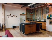 Rustikale Küche mit Holzboden und Wänden aus unverputztem Mauerwerk und Holzrahmen. Zentral steht eine Kücheninsel mit schwarzer Arbeitsplatte; im Hintergrund befinden sich Hängeschränke, ein Waschbecken und eine Arbeitsfläche mit Küchengeräten.