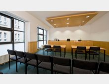 Ein moderner Gerichtssaal mit schlichten Holzmöbeln, einem erhöhten Richterpult und Reihen von schwarzen Stühlen für Zuschauer. Große Fenster an den Seiten lassen Tageslicht herein, und die weiße Decke enthält runde Deckenspots.