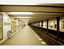 U-Bahn-Station mit zwei gegenüberliegenden Bahnsteigen und einem Gleis in der Mitte. Die Wände sind mit weißen Fliesen verkleidet, während der Ausgang "Sophie-Charlotte-Platz" auf einem Schild angezeigt wird; vereinzelte Passanten warten auf den Bahnsteigen.